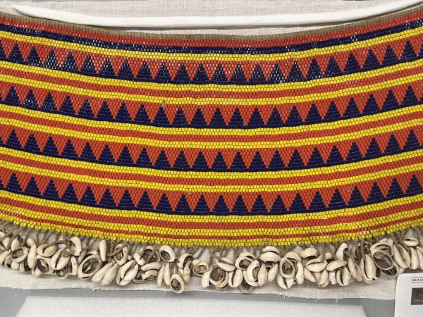 A dibul kouana (woman's cache-sexe) made by an unidentified 20th century Bana Guili artist in Cameroon. Alternating bands of red, navy blue, and yellow beads mix with beaded triangles in navy and red. The bottom edge features cowrie shells.