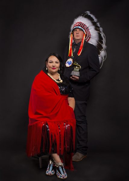 A photograph of Norma Baker-Flying Horse (Red Berry Woman) sitting on the left in a red shawl and Joe Pekara (Pharaoh 171) standing on the right in a traditional headdress.