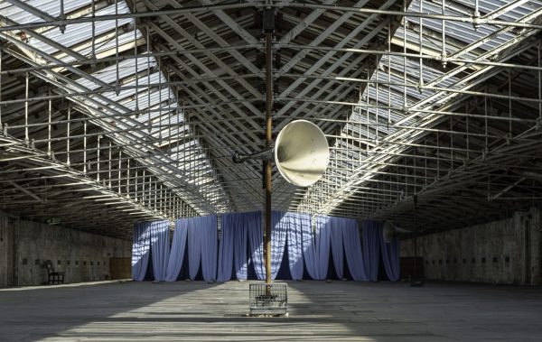 An image of a large scale art installation in a industrial building with a blue curtain in the background and large speakers on poles.